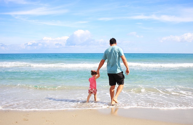 A parent holding their childs hand walk in the shallow waves at the edge of a beach